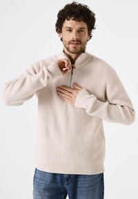 Helles beige, geripptes Strickpullover mit halbem Reißverschlusskragen, langen Ärmeln und lockerer Passform, getragen über blauen Jeans.