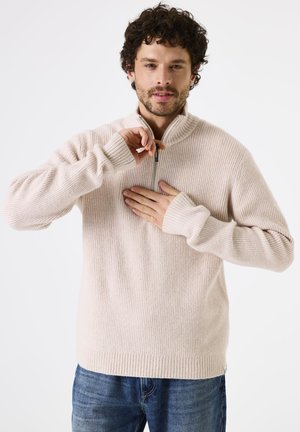 Lichtbeige geribbeld gebreid trui met een halve ritssluiting, voorzien van lange mouwen en een relaxte pasvorm, gedragen over een blauwe jeans.