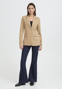 Blazer beige con un solo bottone, revers a punta e due tasche frontali, abbinato a jeans svasati scuri e scarpe nere. Sfondo bianco.