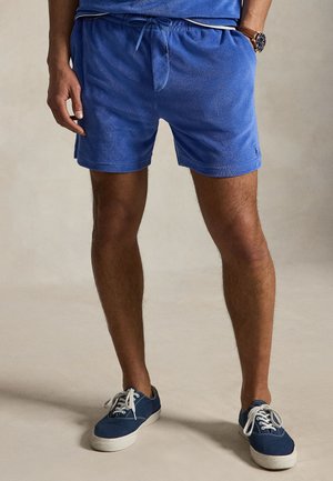 Mand iført blå terry-klæde shorts med snøre, blå lærredsneakers med hvide snørebånd og et armbåndsur på venstre håndled.