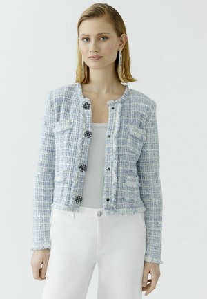 Light blue tweed jacket with frayed edges, five black flower-shaped buttons, and two chest pockets. Worn over a white top and white pants.