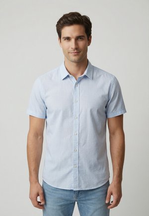 Mann steht vor einem einfachen grauen Hintergrund und trägt ein hellblaues kurzärmeliges Hemd mit Knöpfen und blaue Jeans.