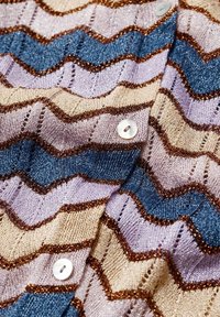 Cardigan en tricot avec un motif en zigzag en beige, lilas et bleu, agrémenté de fils métalliques et de boutons blancs.