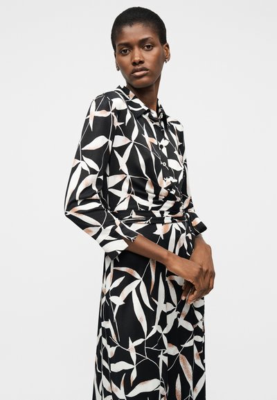 Γυναίκα που φοράει μαύρο φόρεμα με μακριά μανίκια και λευκά και ανοιχτό καφέ αφηρημένα σχέδια φύλλων, στέκεται μπροστά σε ένα απλό ανοιχτόχρωμο φόντο.