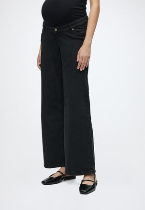 Personne enceinte portant un jean de maternité noir à jambes larges et des chaussures plates noires, debout devant un fond clair uni.