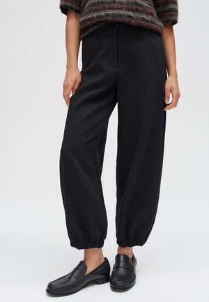 Persona che indossa pantaloni neri con orlo elastico, mocassini in pelle nera e un maglione a righe di lana con le maniche arrotolate.