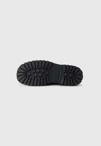 Suela de bota de goma negra con un patrón de tracción texturizado, con agarraderas elevadas, tacón redondeado y surcos profundos para mayor tracción.