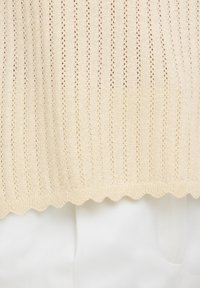 Tissu beige tricoté avec des rayures verticales en ajouré et un bord festonné, posé sur un tissu blanc lisse en dessous.
