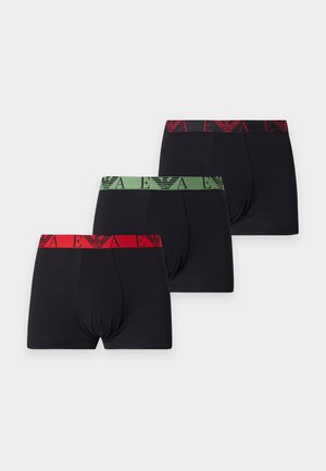 Hombre musculoso de piel oscura, de pie, descalzo, vistiendo bóxers negros con una cintura roja estampada sobre un fondo blanco liso.
