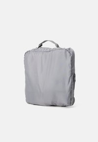 Sac de voyage gris rembourré avec une poignée, présentant une texture lisse et une fermeture zippée sur un côté, compact et de forme carrée.