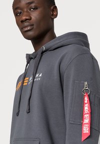 Felpa con cappuccio grigia con tasca frontale, logo arancione sul petto, e patch di tessuto con etichetta rossa "RIMUOVI PRIMA DEL VOLO" sulla manica.