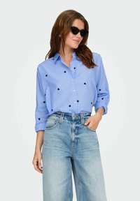 Šviesiai mėlynas dryžuotas marškinis su tamsiai mėlynomis širdelėmis; derinamas su aukštu liemeniu šviesiai mėlynomis plačiomis kelnėmis. Juodos saulės akiniai.