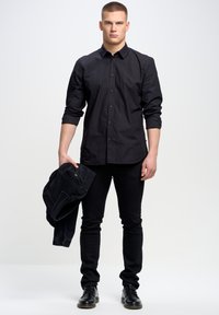 Camisa negra de botones con mangas largas, combinada con jeans negros ajustados y zapatos. Sostiene una chaqueta de mezclilla negra en su mano derecha.
