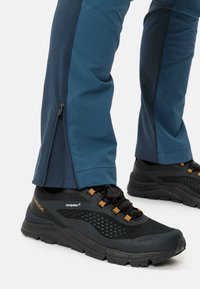 Zwarte wandelschoenen met gele accenten en netpanelen, gecombineerd met technische blauwe broeken met een rits bij de enkels voor een verstelbare pasvorm.