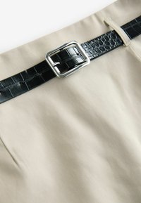 Cintura de tela beige con un cinturón de cuero sintético negro texturizado que presenta una hebilla rectangular plateada, destacando los materiales y colores contrastantes.