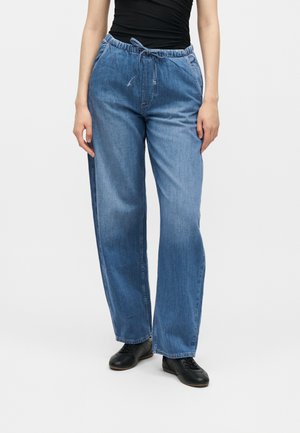 Γυναίκα που φοράει άνετο μπλε τζιν με κορδόνι στη μέση, μαύρο αμάνικο τοπ και μαύρα δερμάτινα παπούτσια με κορδόνια, με μονόχρωμο φόντο.