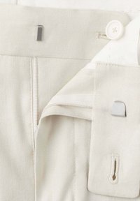 Gros plan sur la ceinture d’un pantalon de couleur crème avec bouton, fermeture éclair, fermeture à crochet et barre, et détails de boucle en tissu.
