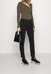 Jeans negros de corte ajustado, combinados con un top de manga larga de canalé en un tono verde oliva oscuro con un corte asimétrico. Sosteniendo un pequeño bolso negro.
