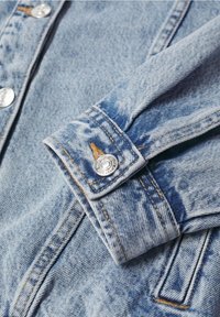 Manche d'une veste en denim bleu clair avec des coutures visibles, un bouton pression argenté, et des détails de surpiqûres orange sur le poignet et le panneau avant.