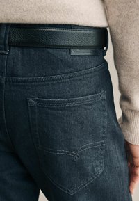 Jeans de mezclilla oscura con un cinturón de cuero negro texturizado. Presentan un diseño distintivo en el bolsillo trasero con detalle de costura en contraste.