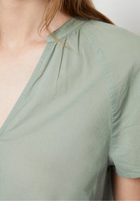 Primo piano di una persona che indossa una blusa verde chiaro con scollatura a V e dettagli in tessuto arricciato vicino alla cucitura della spalla.