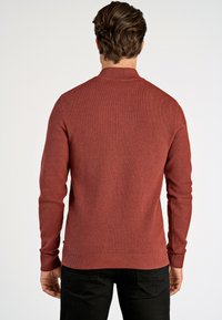 Punainen teksturoitu neulepaita, jossa on korkea kaula-aukko, pitkät hihat sekä ribattuja resoreita hihansuissa ja helmassa, yhdistetty mustiin housuihin, nähtynä takaa.