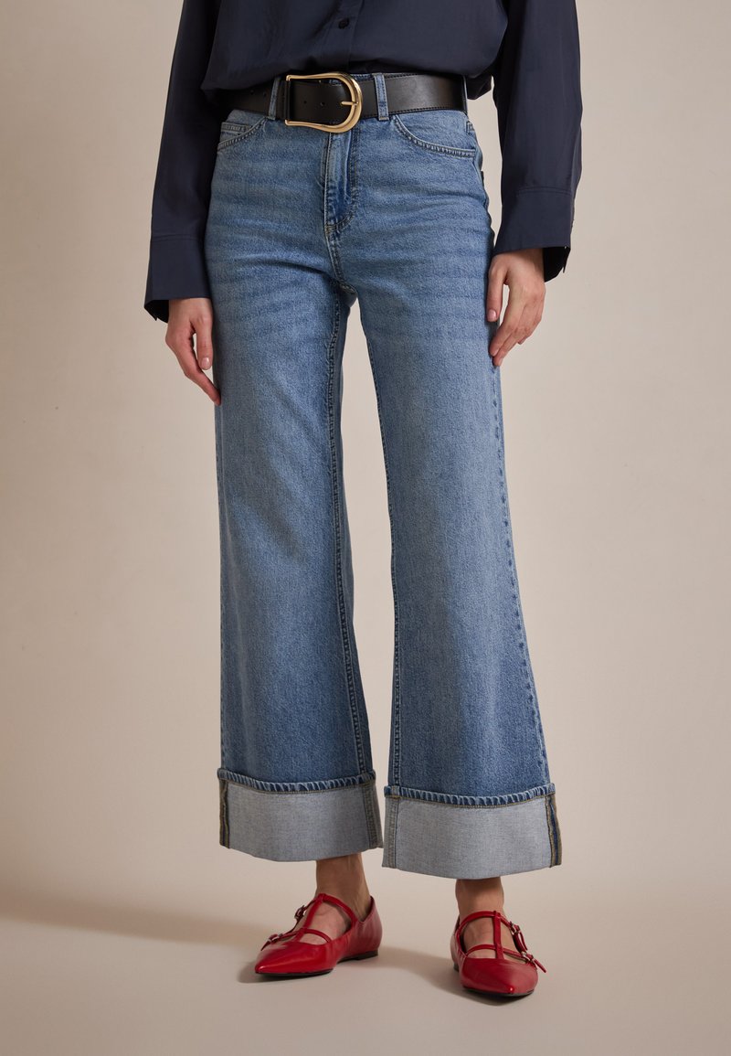 Široke svetlo modre jeans hlače s zavitimi manšetami, v kombinaciji s temno modro srajco in rdečimi lakiranimi čevlji, zavarovanimi z črnim pasom.