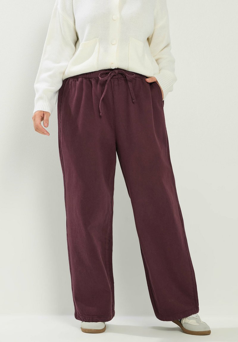 Persoon draagt wijde bordeauxrode broek met trekkoord in de taille, wit gebreid vest met knopen en witte sneakers, staand tegen een effen achtergrond.