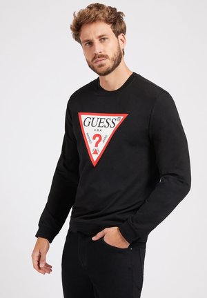 Svart hettegenser med lange ermer, med et rødt trekantet grafikk med hvit tekst "GUESS" og et spørsmålstegn i midten.