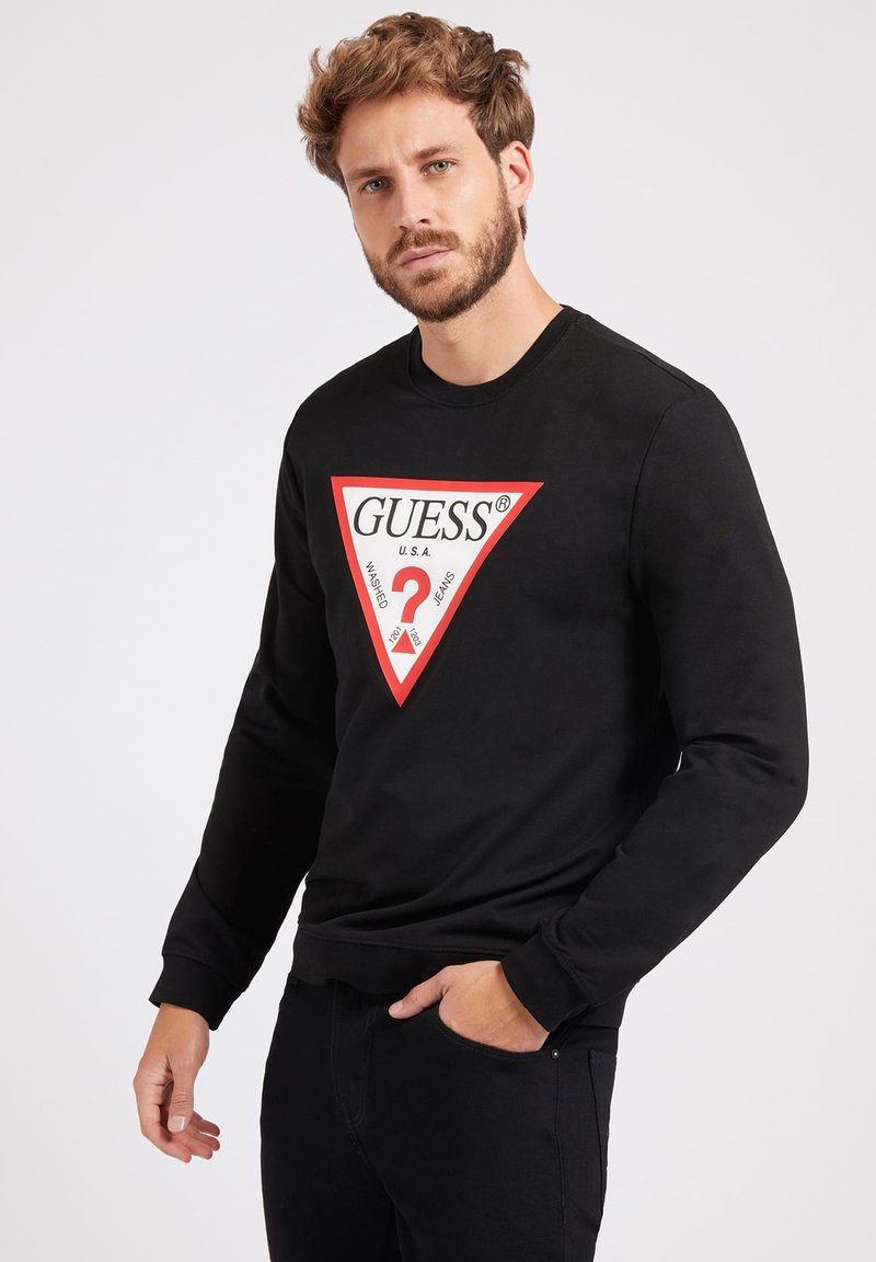 Svart hettegenser med lange ermer, med et rødt trekantet grafikk med hvit tekst "GUESS" og et spørsmålstegn i midten.