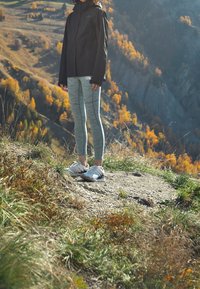 Svart vattentät jacka, grårandiga leggings och vita sportskor på en grusväg med höstlöv i bakgrunden.