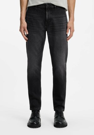 Mann trägt eng anliegende schwarze Jeans und schwarze Schnürschuhe und steht vor einem schlichten weißen Hintergrund.