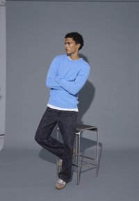 Világoskék kötött pulóver, fekete farmernadrág és szürke cipő. Álló pózban a szürke háttér előtt, mellette egy fém székkel.