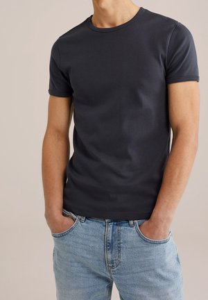 Mężczyzna w gładkiej, granatowej koszulce z krótkim rękawem i jasnoniebieskich jeansach, z rękami w kieszeniach.