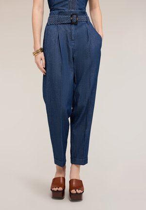 Nainen, joka käyttää korkeavyötäröisiä sinisiä farkkuja punotulla vyöllä, toinen käsi taskussa, yhdistettynä ruskeisiin platform-avokkaisiin ja kultaisiin rannekoruihin.