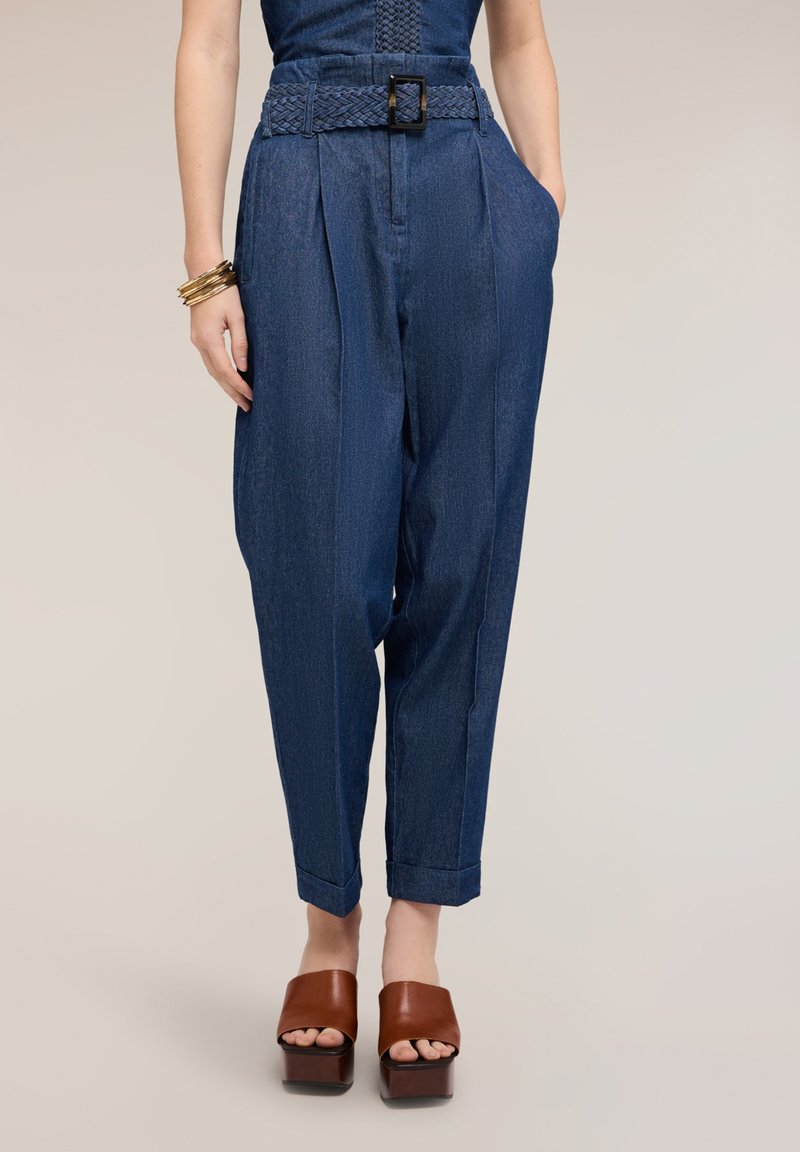 Mujer con pantalones vaqueros azul de talle alto con cinturón trenzado, una mano en el bolsillo, combinados con sandalias marrones de plataforma y pulseras doradas.