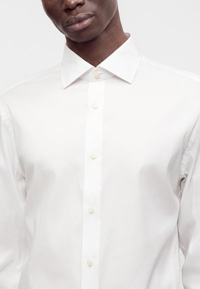 Primo piano di una persona che indossa una camicia bianca impeccabile con colletto appuntito su uno sfondo chiaro e uniforme.