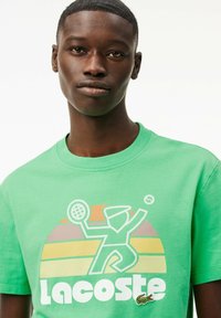 Camiseta de algodón verde con un gran gráfico de un jugador de tenis sosteniendo una raqueta, rayas de atardecer en capas y un pequeño logo de cocodrilo.