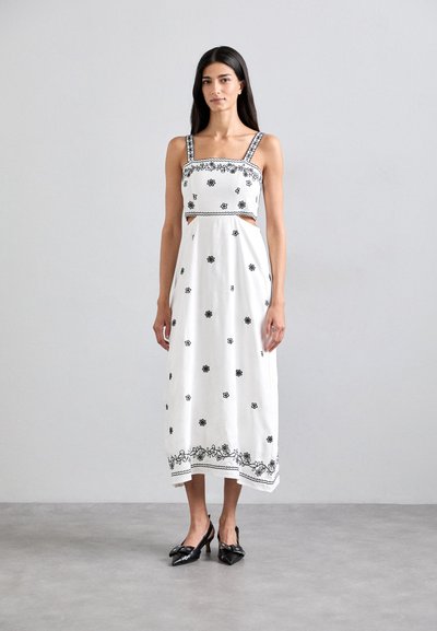 Robe maxi blanche avec broderie de fleurs noires, détail de taille découpé et bretelles ornées. Associée à des chaussures à talons noires.