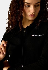 Schwarze Zip-Hoodie aus weichem Stoff mit einem kleinen, weißen, gestickten "Champion"-Logo und einem rot-weißen Akzent am Reißverschluss.