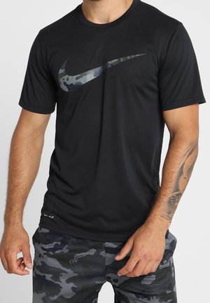 Mann trägt schwarzes Nike Dri-FIT T-Shirt mit grauem Tarnmuster-Swoosh-Logo und graue Tarnmuster-Shorts, sichtbares Unterarmtattoo am rechten Arm.