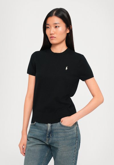 Woman with long dark hair wearing a black short-sleeve shirt with small white logo and blue jeans, hand in pocket, against a plain background.