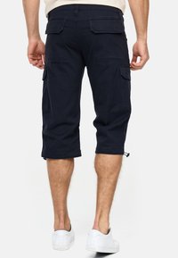 Navy cargo shorts with knee-length design, multiple side pockets, and drawstring hems. Made from durable cotton fabric. White sneakers visible.