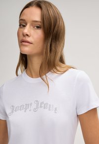 Witte katoenen t-shirt met korte mouwen, versierd met zilveren rhinestones in een scriptlettertype over de borst. Soepele textuur.