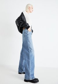 Persona rubia con chaqueta de cuero negra sobre un suéter blanco, jeans sueltos de tiro alto azules y zapatos de plataforma negros, de pie de perfil.