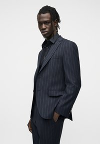 Jeune homme aux dreadlocks portant un costume bleu marine à rayures fines et une chemise noire, debout avec une main dans la poche devant un fond uni.