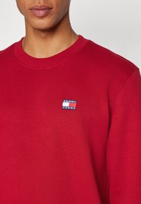 Sudadera roja con cuello redondo acanalado. Presenta un pequeño parche rectangular con el logo en azul, rojo y blanco en el área del pecho izquierdo.