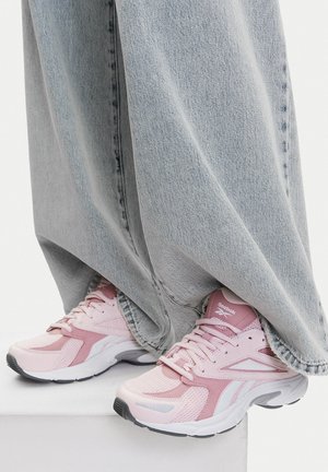 Roza tekaška obuvala z mrežastim in sintetičnim premazom, z belim poudarkom in oblazinjenim podplatom, v kombinaciji z svetlimi kavbojkami.