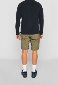 Mies, joka on pukeutunut mustaan, pitkähihainen paita, oliivinvihreät cargo-shortsit, valkoiset sukat ja mustat vaelluskengät, seisoo selin harmaalla lattialla.