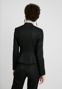 Zwarte op maat gemaakte peplumjacket met lange mouwen, gemêleerde stof en een hoge kraag. Draagt over een grijze coltrui, gecombineerd met bijpassende broek.